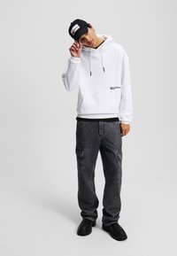 Weißer Kapuzenpullover mit frontalem Taschen- und Zugbanddesign, kombiniert mit dunkelgrauen Cargo-Hosen mit Seitentaschen und schwarzem Schuhwerk.