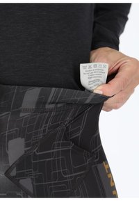 Black leggings featuring a geometric pattern in dark gray, with a visible care label inside the waistband and a zippered pocket.