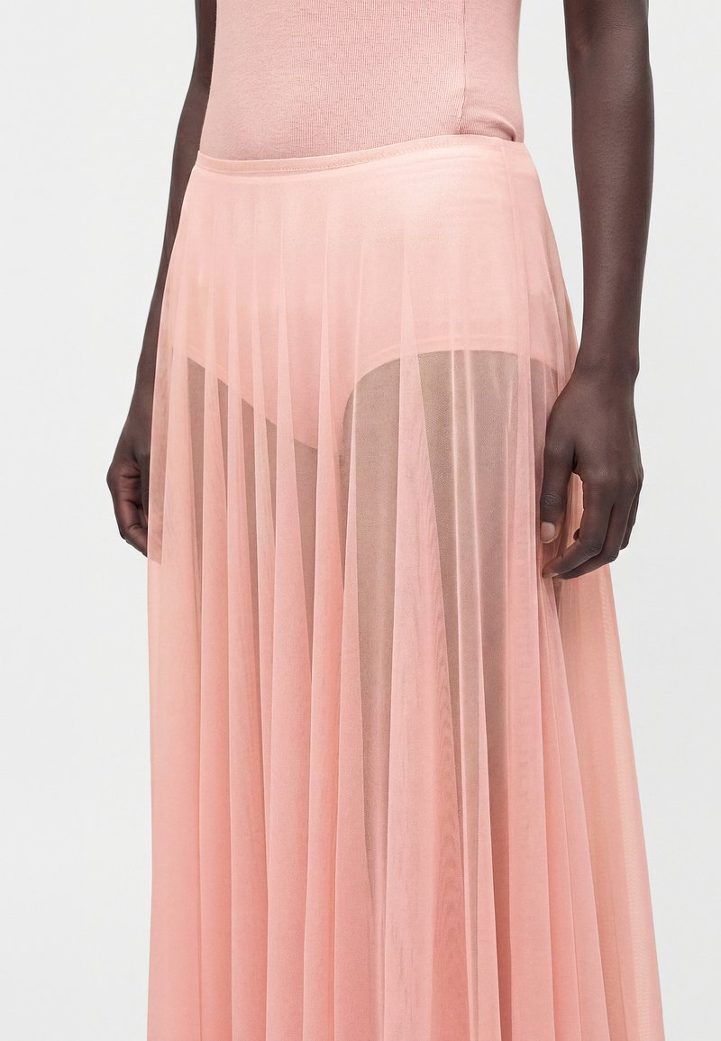Personne à la peau foncée portant une jupe rose plissée transparente révélant des sous-vêtements assortis et un haut rose ajusté, debout devant un fond uni.