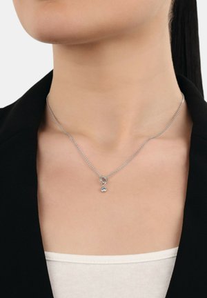 Collar de cadena de plata con un pequeño colgante redondo, llevado en el cuello de una mujer vestida con una chaqueta negra y una blusa blanca.