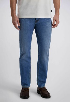 Man met lichtblauwe spijkerbroek, wit T-shirt en donkerbruine leren loafers, staande tegen een effen grijze achtergrond.