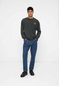 Langermet mørkegrå bomullsskjorte med en subtil logo, kombinert med blå denimjeans og sorte sneakers. En enkel og uformell design.