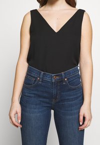 Svart ärmlös topp med djup V-ringning, tillverkad av mjuk tyg. Matchas med högmidjade mörkblå jeans med koppardetaljer.