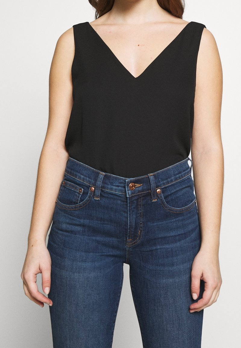 Svart ärmlös topp med djup V-ringning, tillverkad av mjuk tyg. Matchas med högmidjade mörkblå jeans med koppardetaljer.