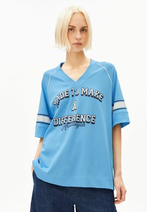 Jeune personne aux cheveux blonds courts porte une chemise bleue ample avec le texte "Made to Make a Difference" et un pantalon bleu foncé.