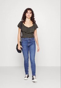 Top rufado verde escuro, jeans azuis justos, mala tiracolo preta e ténis pretos e brancos. Design suave e casual com um ajuste relaxado.