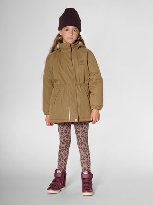Fille portant une parka marron à capuche, un bonnet violet, des leggings imprimés léopard et des baskets montantes violettes, debout devant un fond gris uni.