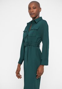 Tumeroheline pikkade varrukatega kleit, millel on kaks esist taskut, vööga talje ja struktureeritud krae. Siledat tekstuuri.