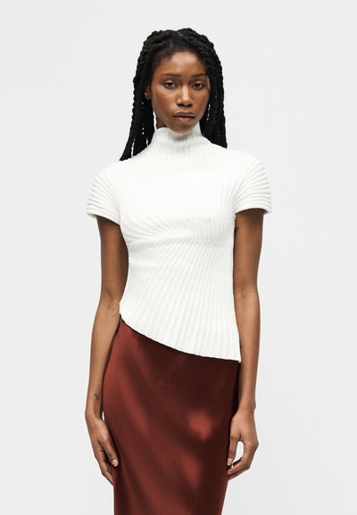 Femme aux longues tresses portant un haut à manches courtes côtelé blanc à col roulé et une jupe en satin marron, debout devant un fond neutre.
