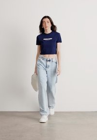 Jūrinis trumpas marškinėliai su balta užrašu, šviesiai mėlyni aukšto liemens džinsai ir baltos sportbačiai. Modelis neša šviesią rankinę, eidamas neutraliame fone.