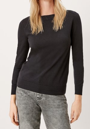Vrouw draagt een aansluitende zwarte trui met lange mouwen en lichtgrijze jeans met een geëtste wassing, staand tegen een effen lichte achtergrond.