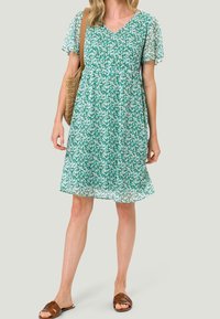 Vestido floral verde con escote en V, mangas cortas con volantes, tejido ligero y corte holgado. Combinado con un bolso marrón y sandalias.