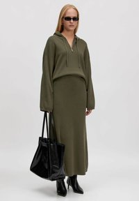 Olijfgroen sweater jurk met lange mouwen en een ritskap, gecombineerd met zwarte hoge hakken en een glanzende zwarte tote tas.