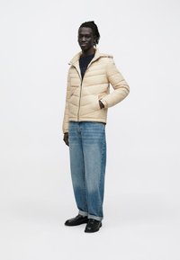 Μπεζ μπουφάν με υφή από ραμμένα τμήματα και κλείσιμο με φερμουάρ, συνδυασμένο με μπλε τζιν παντελόνι και μαύρα παπούτσια. Το μοντέλο στέκεται με τα χέρια στις τσέπες.