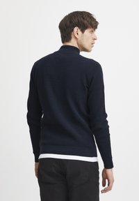 Pulover tricotat bleu închis, cu guler înalt, țesătură texturată și manșete cu model, purtat peste o cămașă albă, alături de pantaloni negri.