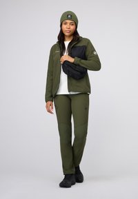 Olive green fleece jacket with black chest panel, matching pants, and a green beanie. Black bag worn across the body. Lightweight fabric.
