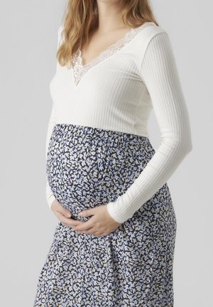 Kobieta w ciąży ubrana w biały top z długim rękawem i koronkowym detalem oraz niebieską kwiecistą spódnicę, delikatnie obejmująca brzuch.