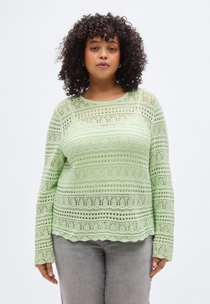 Jasnozielony sweter z długim rękawem o fakturowanym wzorze dzianiny, zawierający różne motywy i falbankę na dole, w połączeniu z szarymi spodniami.