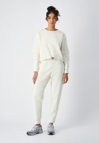 Sudadera de crema claro con un diseño recortado, dobladillo elástico y mangas largas, combinada con pantalones ajustados a juego y zapatillas deportivas grises.