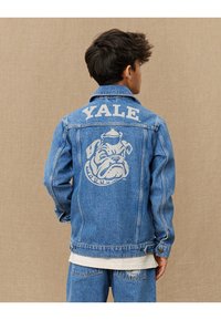 Veste en denim bleu clair avec un imprimé blanc "YALE" et un graphique de bulldog au dos. Dotée d'un col classique et d'une fermeture à boutons sur le devant.