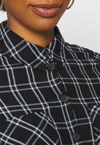 Svartvit rutigt skjorta med krage, knappstängning och texturerad tyg. Innehåller subtila blå accenter i mönstret.