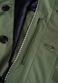 Prenda exterior verde con botones negros, que cuenta con un bolsillo con cremallera y un ribete a cuadros en contraste a lo largo de la abertura. Textura suave y lisa.