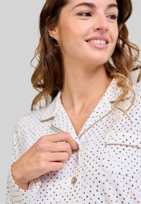Mujer sonriendo y abrochándose una parte superior de pijama blanca con lunares azules y marrones y un bolsillo en el frente, con el cabello rizado visible.