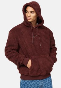 Felpa con cappuccio bordeaux in pile con una consistenza morbida, dettagli con cordino e tasca a marsupio. Mani e fondo a costine. Logo ricamato sulla parte frontale.