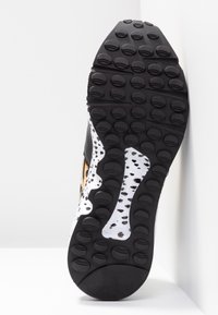 Semelle de baskets noires avec des motifs de semelle circulaires et une section blanche présentant des taches noires, reposant verticalement contre un coin blanc.