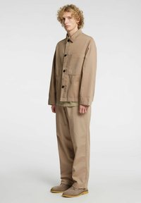 Lysebrun bomullsjakke med lange ermer, frontlomme og svarte knapper. Kombinert med beige bukser og grå støvler, mot en ensfarget bakgrunn.