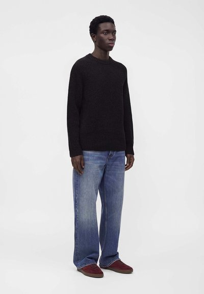 Jeune homme portant un pull noir, un jean bleu ample et des chaussures rouges, debout devant un fond blanc uni.