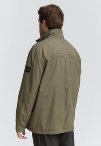 Olivengrüne Jacke aus leichtem Material mit strukturiertem Kragen, langen Ärmeln und schwarzem Logopatch auf der linken Schulter.
