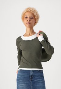 Olíva zöld bordás, hosszú ujjú felső egy fehér póló fölé rétegezve, kék farmer szoknyával párosítva. A modell egy olíva táskával a kezében.