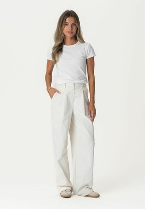 White t-shirt with short sleeves paired with high-waisted white wide-leg pants and light-colored shoes, standing casually with hands in pockets.