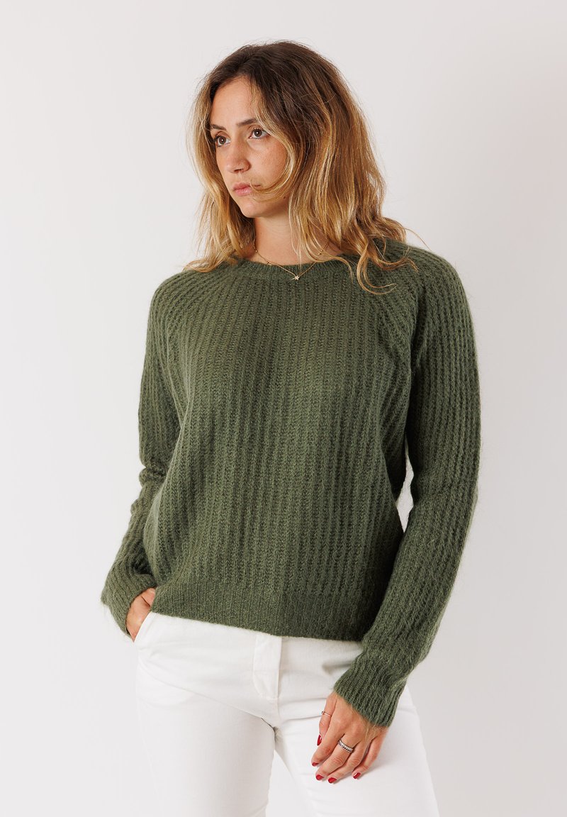 Donna con capelli biondi ondulati, che indossa un maglione a coste verde oliva e pantaloni bianchi, in piedi con una mano in tasca davanti a uno sfondo semplice.