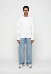 Νέος άνδρας που στέκεται προς τα εμπρός, φορώντας ένα απλό λευκό μακρυμάνικο πουκάμισο, φαρδιά ανοιχτό μπλε τζιν και λευκά αθλητικά παπούτσια πάνω σε λευκό φόντο.