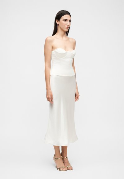 Γυναίκα που φοράει λευκό στράπλες κορσέ και ταιριαστή φούστα μέσης μήκους, με χρυσά ανοιχτά τακούνια, σε απλό φόντο.