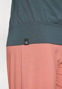 Teal and coral two-piece outfit featuring a gathered waist design. Black fabric tag with logo on the side of the top near the seam.