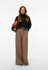 Kobieta w czarnym golfie, szerokich brązowych spodniach z paskiem, trzymająca dużą brązową skórzaną kopertówkę, stojąca na białym tle.