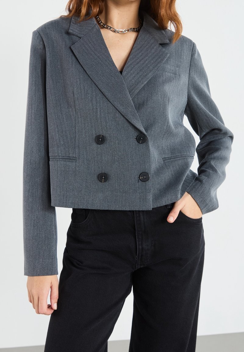 Femme portant un blazer gris court à double boutonnage avec des boutons noirs, un pantalon noir et un collier chaîne argenté, une main dans la poche.