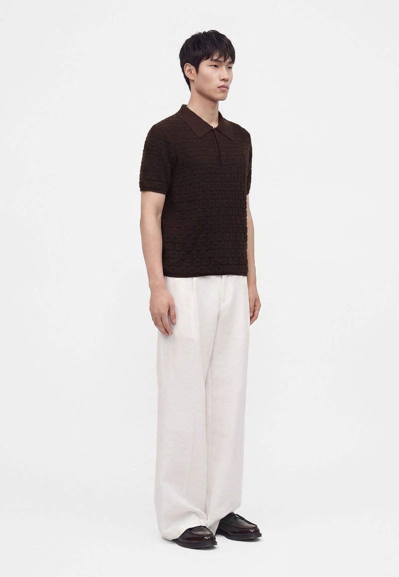 Hombre de pie con una camiseta polo de manga corta oscura y texturizada, pantalones blancos holgados y zapatos oscuros contra un fondo claro y sencillo.
