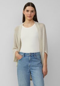 Helle Beige, offen gestrickte Strickjacke mit weiten Ärmeln, getragen über einem weißen, enganliegenden Oberteil und kombiniert mit hellblauen Jeans. Glatte Textur, lockere Passform.