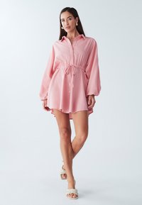 Lichtroze shirtjurk met een kraag, trekkoord in de taille en lange mouwen. Glad materiaal met een iets uitlopende zoom. G gedragen met gevlochten sandaal.