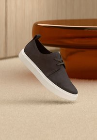 Σκούρο γκρι αθλητικό παπούτσι με κορδόνια και λευκή σόλα να αιωρείται πάνω από μπεζ χαλί, με ένα πορτοκαλί γυαλιστερό στρογγυλό τραπέζι στο φόντο.