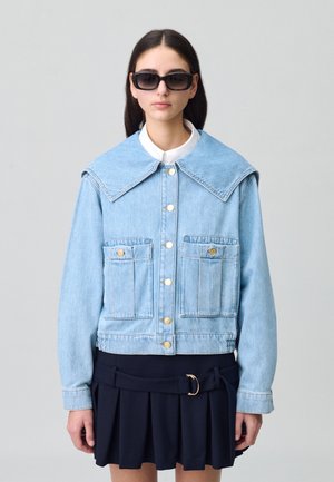 Chaqueta de denim azul claro con gran cuello, botones dorados y dos bolsillos en el pecho, combinada con una falda oscura con pliegues y una camisa blanca con cuello.