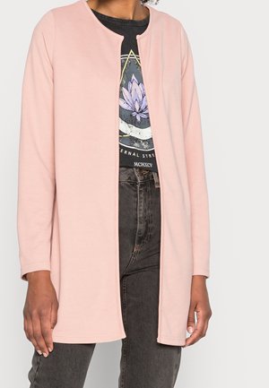 Cardigan long rose en texture côtelée, avec un col rond et un design ouvert à l'avant. Porté par-dessus un T-shirt noir à motif graphique et un jean sombre.
