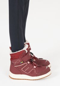 Bottes d'hiver imperméables rouges avec un col rembourré, des lacets et une semelle en caoutchouc, accompagnées de leggings noirs. Couture accentuée et logo visibles.