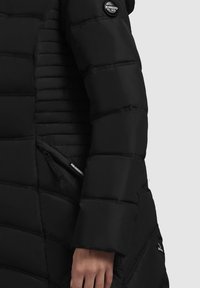 Svart puffjacka med vadderad design, som har en jämn textur, diagonala quiltningar och sidfodrade fickor med dragkedja. Märkespatch på axeln.