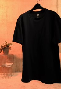 Črna kratka majica GAP visi na črnem obešalniku z žično okvirno stransko mizico in sobno rastlino v lončku na ozadju toplega prostora.