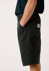 Shorts de mezclilla negra con corte recto, bolsillos laterales y un parche de logo cosido en el bolsillo trasero. Presenta un detalle azul cerca del borde del bolsillo.